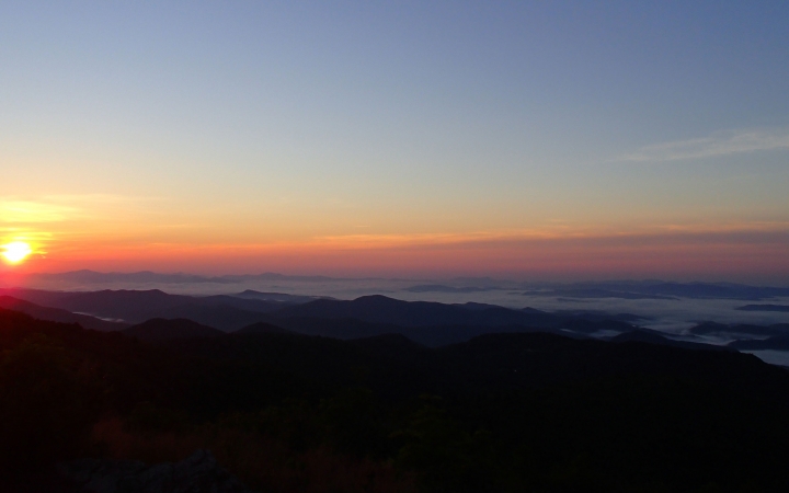 The sun appears to be setting or rising in a mountainous landscape. The sky appears in soft shades of blue, yellow, orange, pink and purple.